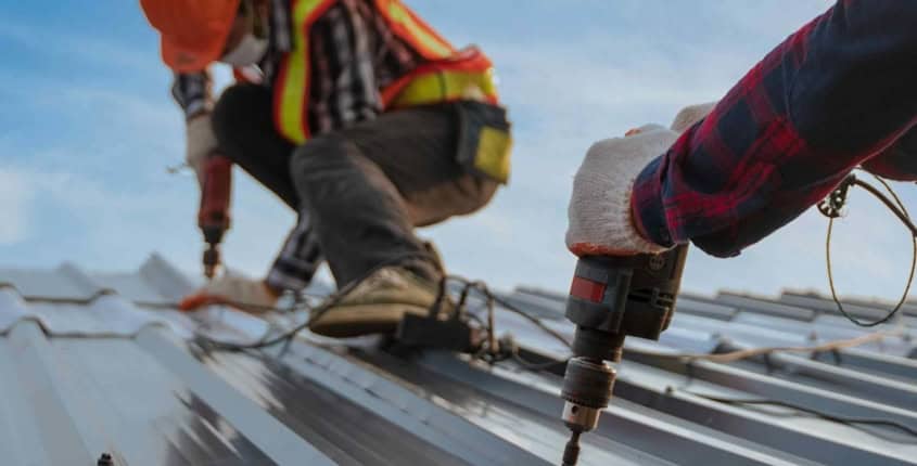 May-Blog-2 Two roofers laying metal on roof surface, using tools
